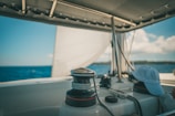 A boat deck with sailing equipment and ropes visible under a shaded canopy. The ocean stretches into the distance with a partly cloudy sky. A white cap is resting on the deck.