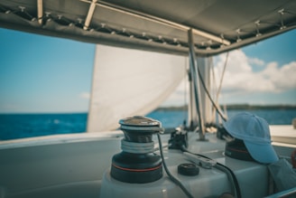 A boat deck with sailing equipment and ropes visible under a shaded canopy. The ocean stretches into the distance with a partly cloudy sky. A white cap is resting on the deck.
