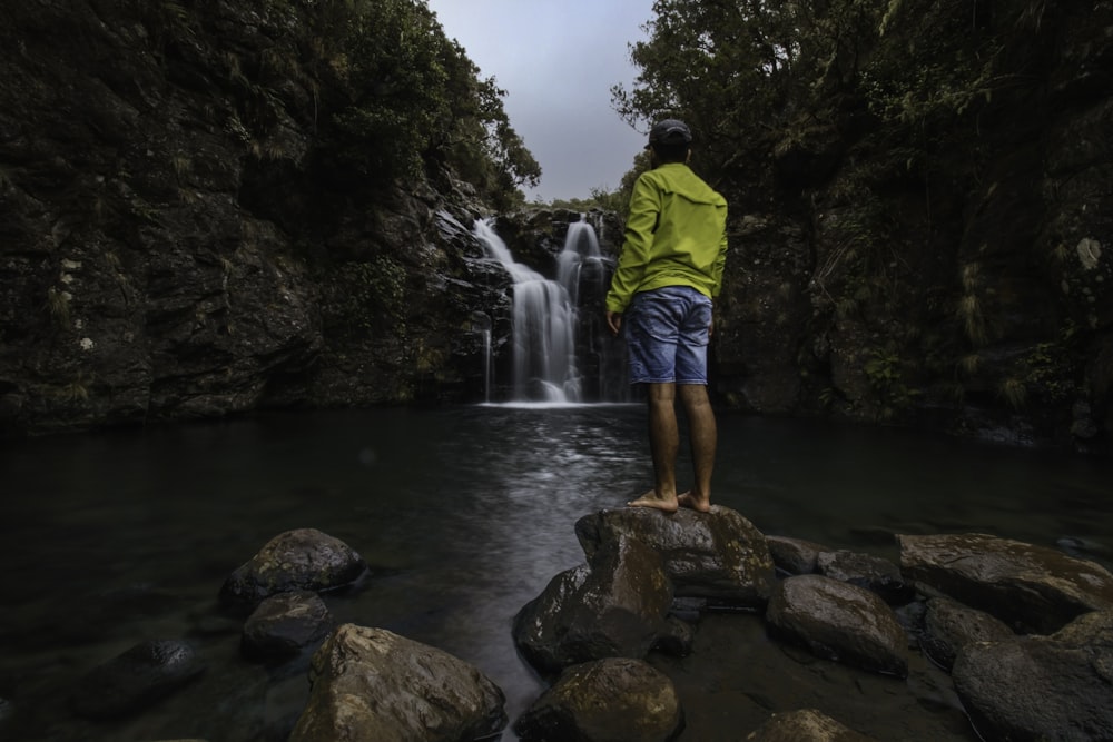 man in yellow shirt and blue shorts standing on brown rock near waterfalls during daytime