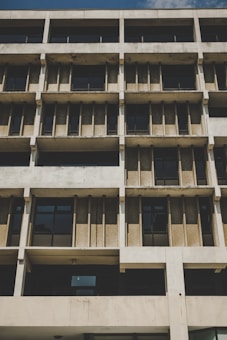 A multi-story building with a Brutalist architectural style features repetitive concrete patterns and large rectangular windows. The facade displays raw concrete surfaces with clear geometric lines and a rough texture.