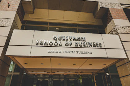 A modern building entrance with a sign reading 'Questrom School of Business Rafik B. Hariri Building' in metallic lettering. The facade features a combination of glass and stone materials, with decorative stone patterns visible. Warm lighting is present, adding a welcoming atmosphere.