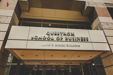 A modern building entrance with a sign reading 'Questrom School of Business Rafik B. Hariri Building' in metallic lettering. The facade features a combination of glass and stone materials, with decorative stone patterns visible. Warm lighting is present, adding a welcoming atmosphere.