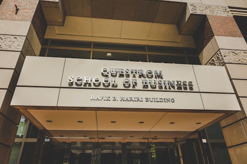 A modern building entrance with a sign reading 'Questrom School of Business Rafik B. Hariri Building' in metallic lettering. The facade features a combination of glass and stone materials, with decorative stone patterns visible. Warm lighting is present, adding a welcoming atmosphere.
