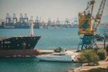 A busy port scene with large cargo ships and a series of cranes in the background. A smaller yacht is docked alongside a larger cargo ship. The water is calm and there are industrial structures along the coastline.