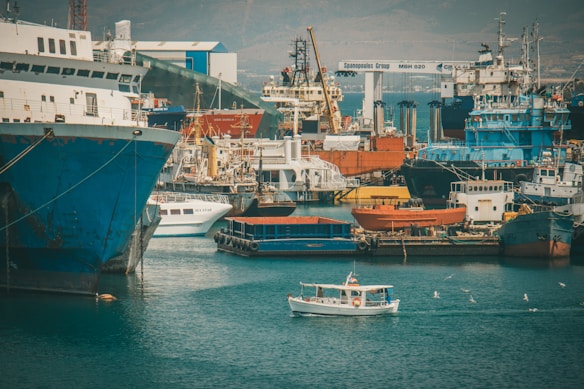 A bustling shipyard with numerous docked vessels, including large cargo ships and a small boat sailing in the foreground. The area is filled with cranes and industrial structures, suggesting an active maritime industrial zone. Gulls are flying over the water, adding a lively touch to the scene.