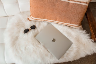 A cozy setup featuring a closed laptop with an Apple logo placed on a fluffy white fur rug. Beside the laptop, there is a pair of dark sunglasses. An orange cushion with white stripes rests on a white cushioned seat.