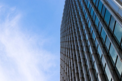 The image captures a modern skyscraper from a low-angle perspective against a bright blue sky. The building features numerous windows reflecting the sky, creating a sense of height and architectural finesse.