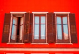 A vibrant red building facade with three adjacent wooden windows that are partially open. The windows have shutters, and elegant wrought iron railings are visible below each window, adding character to the classic architectural design.