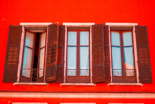 A vibrant red building facade with three adjacent wooden windows that are partially open. The windows have shutters, and elegant wrought iron railings are visible below each window, adding character to the classic architectural design.