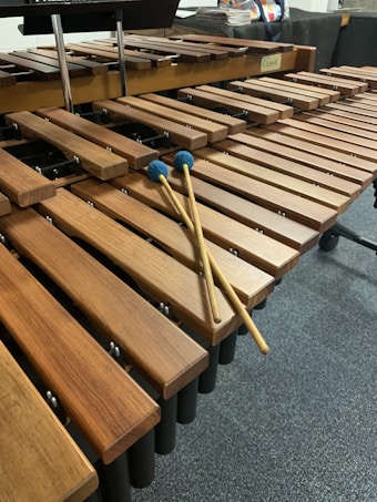 A wooden percussion instrument with several bars arranged in rows, resembling a xylophone. Two mallets with blue heads are resting on the bars. The setting appears to be a music room or studio.