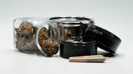 A collection of cannabis-related items, including a glass jar filled with cannabis buds, a metal grinder with its lid off, and a rolled joint laying in front. The scene features earthy tones and metallic highlights.