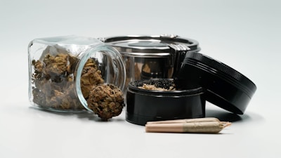 A collection of cannabis-related items, including a glass jar filled with cannabis buds, a metal grinder with its lid off, and a rolled joint laying in front. The scene features earthy tones and metallic highlights.