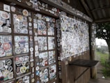 A wooden wall and table surface are covered with hundreds of colorful stickers, showcasing various designs, logos, and motifs. The surface is part of a rustic structure with wooden beams and a corrugated metal roof. The scene is set outdoors, with a glimpse of greenery and overcast skies in the background.