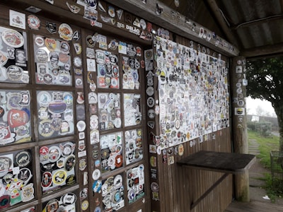 A wooden wall and table surface are covered with hundreds of colorful stickers, showcasing various designs, logos, and motifs. The surface is part of a rustic structure with wooden beams and a corrugated metal roof. The scene is set outdoors, with a glimpse of greenery and overcast skies in the background.