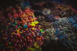 A vibrant assortment of colorful knitted objects and small dolls are displayed on a surface. The collection includes a variety of keychains, bracelets, and figures, arranged in sections by type. The items are illuminated under soft lighting, creating a cozy and inviting marketplace atmosphere.