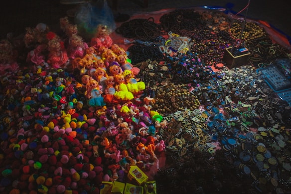 A vibrant assortment of colorful knitted objects and small dolls are displayed on a surface. The collection includes a variety of keychains, bracelets, and figures, arranged in sections by type. The items are illuminated under soft lighting, creating a cozy and inviting marketplace atmosphere.