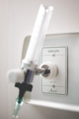 A close-up view of a wall-mounted oxygen outlet in a clinical or hospital setting, featuring a metal connector and a flow meter valve. The panel has marked instructions and the background is a neutral light color.