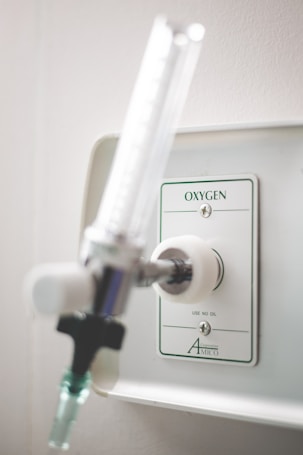 A close-up view of a wall-mounted oxygen outlet in a clinical or hospital setting, featuring a metal connector and a flow meter valve. The panel has marked instructions and the background is a neutral light color.
