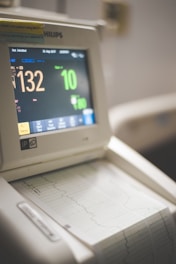 A medical monitor with a digital display shows various patient statistics, including heart rate and other vital signs. The device is placed above a printed graph output, likely representing a heart rate or other medical measurement over time. The screen features bright numeric and graphical displays.