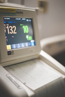 A medical monitor with a digital display shows various patient statistics, including heart rate and other vital signs. The device is placed above a printed graph output, likely representing a heart rate or other medical measurement over time. The screen features bright numeric and graphical displays.