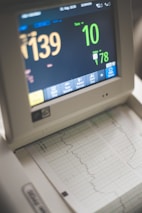 A medical monitor displaying various vital signs including heart rate and pulse. The screen shows numbers like 139 and 78, with graphical representations of the data below on a printout. The device has several buttons for interaction at the bottom.