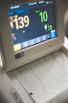 A medical monitor displaying various vital signs including heart rate and pulse. The screen shows numbers like 139 and 78, with graphical representations of the data below on a printout. The device has several buttons for interaction at the bottom.