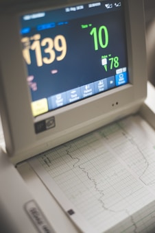 A medical monitor displaying various vital signs including heart rate and pulse. The screen shows numbers like 139 and 78, with graphical representations of the data below on a printout. The device has several buttons for interaction at the bottom.