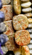 A close-up view of polished stones or minerals, featuring a range of earthy tones such as brown, green, and orange. The stones have a smooth, shiny finish, and some exhibit a marbled pattern.