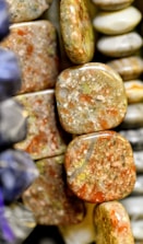 A close-up view of polished stones or minerals, featuring a range of earthy tones such as brown, green, and orange. The stones have a smooth, shiny finish, and some exhibit a marbled pattern.