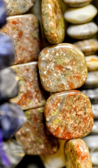 A close-up view of polished stones or minerals, featuring a range of earthy tones such as brown, green, and orange. The stones have a smooth, shiny finish, and some exhibit a marbled pattern.