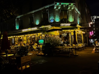 A lively outdoor cafe scene at night, with people sitting at tables under warm, glowing lights. The building has decorative lighting that highlights its architectural features, creating a cozy and inviting atmosphere. The sidewalk features potted plants and umbrellas, adding to the ambiance.