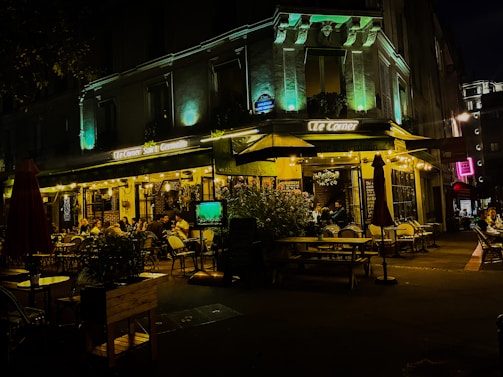 A lively outdoor cafe scene at night, with people sitting at tables under warm, glowing lights. The building has decorative lighting that highlights its architectural features, creating a cozy and inviting atmosphere. The sidewalk features potted plants and umbrellas, adding to the ambiance.