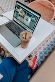A person sitting at a table next to an open laptop displaying a photo editing software. A glass of iced coffee with a straw sits on a napkin beside the laptop. The person is wearing blue pants and red sneakers, and there is a geometric patterned floor and chair visible in the background.