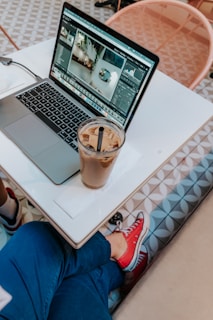 A person sitting at a table next to an open laptop displaying a photo editing software. A glass of iced coffee with a straw sits on a napkin beside the laptop. The person is wearing blue pants and red sneakers, and there is a geometric patterned floor and chair visible in the background.