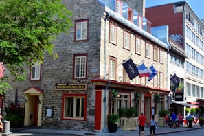 A picturesque street scene featuring a stone building with red-trimmed windows and multiple flags hanging from the entrance, including a French and a Quebec flag. Pedestrians are walking by, and a leafy tree partially shades the sidewalk.