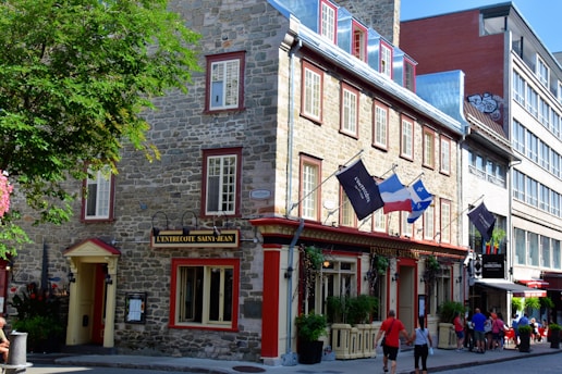 A picturesque street scene featuring a stone building with red-trimmed windows and multiple flags hanging from the entrance, including a French and a Quebec flag. Pedestrians are walking by, and a leafy tree partially shades the sidewalk.