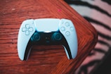 A video game controller lies on a wooden table. The controller is primarily white with black accents and features various buttons and joysticks, characteristic of a gaming accessory.