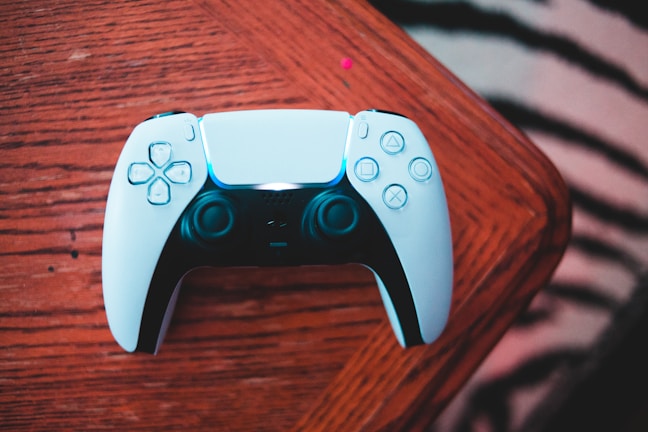 A video game controller lies on a wooden table. The controller is primarily white with black accents and features various buttons and joysticks, characteristic of a gaming accessory.