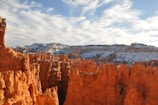 A landscape featuring striking red rock formations known as hoodoos, with a backdrop of snow-dusted mountains beneath a sky filled with scattered clouds.