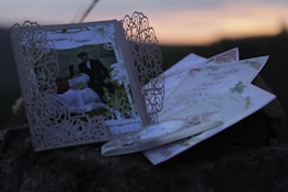 An intricately designed wedding invitation with a laser-cut floral pattern is prominently displayed against a blurred outdoor background. The invitation features a photo of a couple dressed in formal attire. Beside it, there are several other decorative cards and envelopes, adding elegance and sophistication to the arrangement.