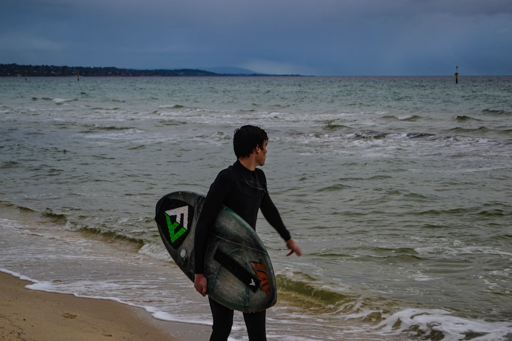 homme en veste noire tenant une planche de surf noire sur la plage pendant la journée