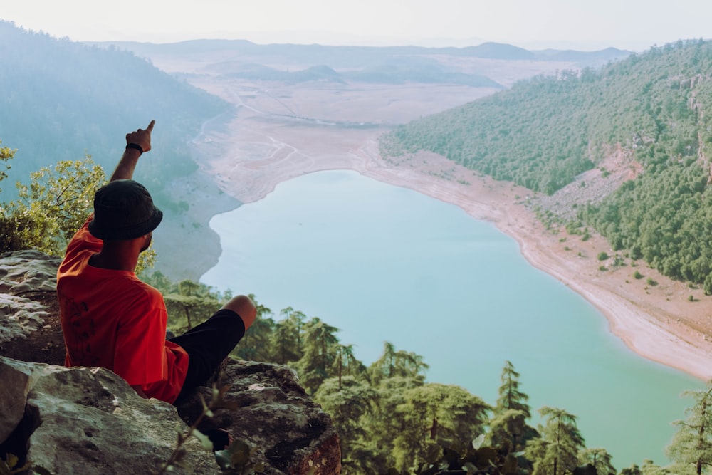 man in red shirt sitting on rock formation looking at lake during daytime