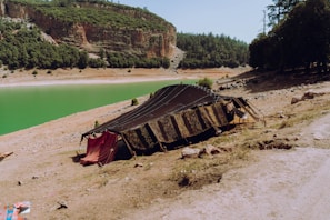 A traditional tent-like structure is set up on a grassy and rocky slope near a serene green lake. The landscape is surrounded by dense forested hills and a steep cliff in the background, creating a picturesque and isolated setting.
