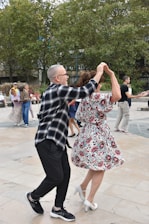 A couple dances energetically in an outdoor setting, surrounded by others either dancing or watching. The man wears a black and white checkered shirt with black pants, and the woman is in a floral dress with white heels. They appear to be enjoying the moment with enthusiasm. The background features trees and several onlookers.