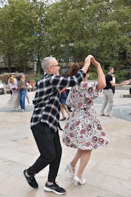 A couple dances energetically in an outdoor setting, surrounded by others either dancing or watching. The man wears a black and white checkered shirt with black pants, and the woman is in a floral dress with white heels. They appear to be enjoying the moment with enthusiasm. The background features trees and several onlookers.