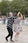 A couple dances energetically in an outdoor setting, surrounded by others either dancing or watching. The man wears a black and white checkered shirt with black pants, and the woman is in a floral dress with white heels. They appear to be enjoying the moment with enthusiasm. The background features trees and several onlookers.