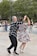 A couple dances energetically in an outdoor setting, surrounded by others either dancing or watching. The man wears a black and white checkered shirt with black pants, and the woman is in a floral dress with white heels. They appear to be enjoying the moment with enthusiasm. The background features trees and several onlookers.