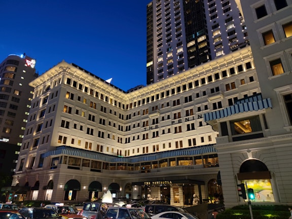 A grand, multi-story hotel building with a classic architectural design stands illuminated against the evening sky. The building features numerous windows with light shining from some, creating a warm ambiance. Blue and white striped awnings adorn parts of the facade. In front of the building, a line of cars is visible, suggesting a busy urban setting.