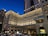 A grand, multi-story hotel building with a classic architectural design stands illuminated against the evening sky. The building features numerous windows with light shining from some, creating a warm ambiance. Blue and white striped awnings adorn parts of the facade. In front of the building, a line of cars is visible, suggesting a busy urban setting.