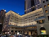 A grand, multi-story hotel building with a classic architectural design stands illuminated against the evening sky. The building features numerous windows with light shining from some, creating a warm ambiance. Blue and white striped awnings adorn parts of the facade. In front of the building, a line of cars is visible, suggesting a busy urban setting.